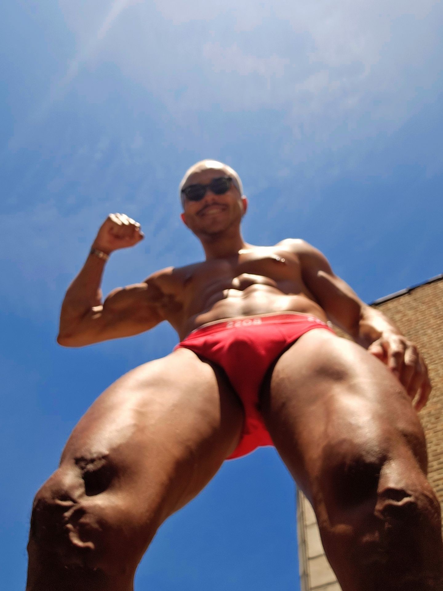 Muscular person in red swimwear posing outdoors with a blue sky background.